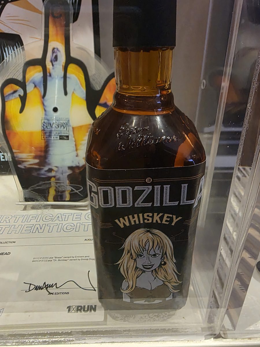 A Godzilla Whiskey bottle with a blonde cartoon woman on the label, displayed in an acrylic case at The Trailer alongside Slim Shady artwork and a 1xRUN certificate of authenticity