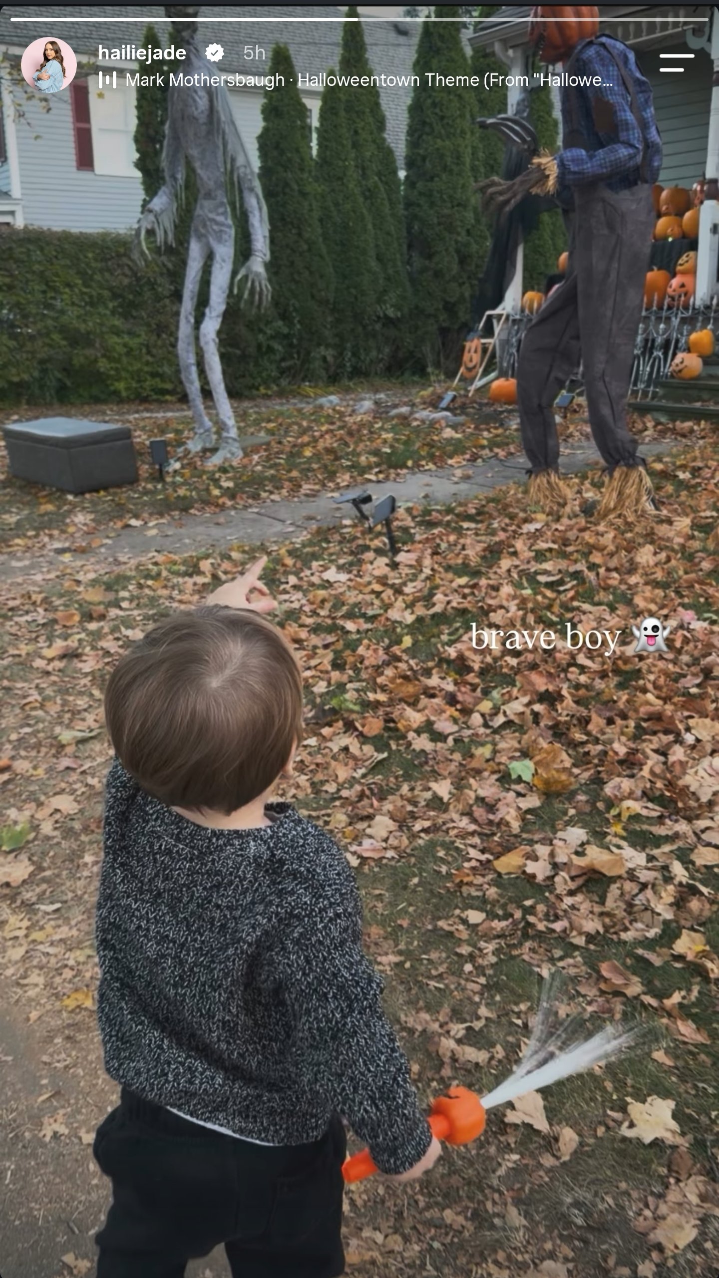 Hailie Jade's young son in a dark knit sweater, holding an orange light-up toy and pointing at a tall skeletal figure and a pumpkin-head scarecrow on a leaf-covered lawn, captioned brave boy