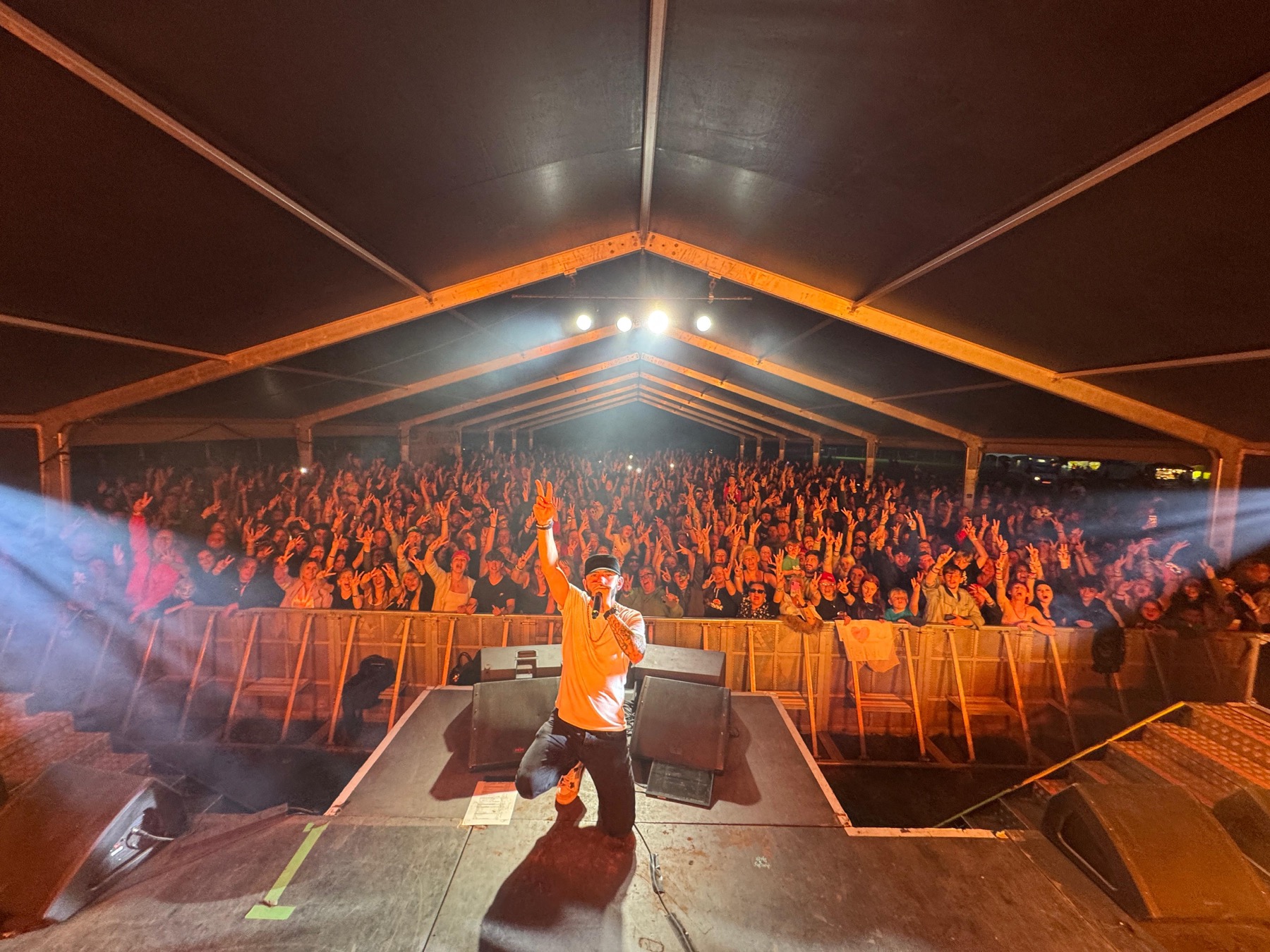 Michael Mathers performing his Eminem tribute set at Festwich, Heaton Park, Manchester in 2025 — shot from the stage behind him, mic in hand and one arm raised, in a white tee, black cap and jeans, facing a packed marquee crowd with hands in the air behind the barrier