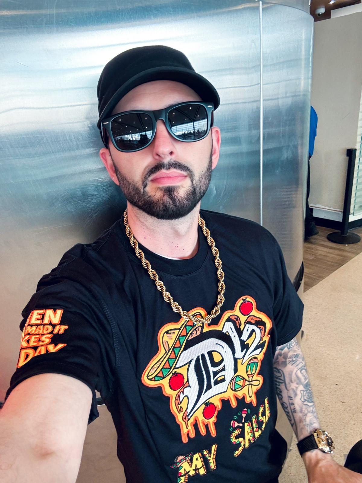 Michael Mathers in a black D12 “My Salsa” T-shirt featuring the D12 Old English logo over a dripping salsa graphic with maracas and a sombrero, wearing a black cap, dark sunglasses, a gold rope chain and a gold watch
