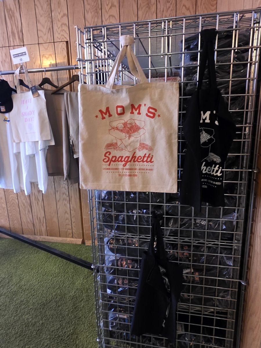 A canvas Mom's Spaghetti tote bag with red spaghetti carton artwork hanging on a wire merch rack inside The Trailer shop, with wood panelling and green carpet